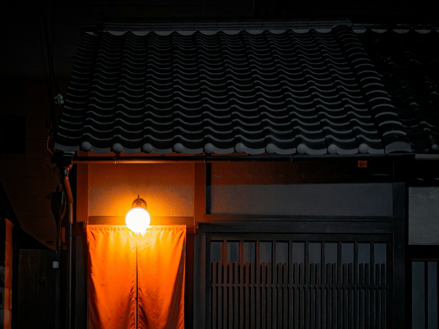 夜の店先。瓦屋根の下、灯りひとつに照らされた橙の暖簾。